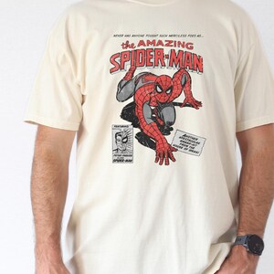K&ouml;nnte beinhalten: Elfenbeinfarbenes T-Shirt mit einem Comic-Motiv von Spider-Man. Das Design enth&auml;lt den Text "the AMAZING SPIDER-MAN" in Rot und Wei&szlig; sowie weitere Texte und Illustrationen. Das T-Shirt hat kurze &Auml;rmel.