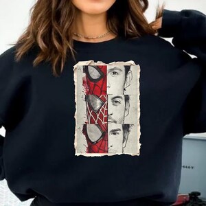Op de afbeelding: Zwarte sweatshirt met een vintage-stijl grafische afbeelding. De afbeelding toont het masker van Spider-Man en de gezichten van drie acteurs die het personage hebben gespeeld. Het ontwerp gebruikt rood, wit en zwart.