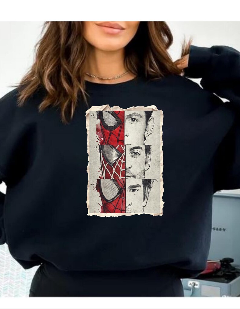 Op de afbeelding: Zwarte sweatshirt met een vintage-stijl grafische afbeelding. De afbeelding toont het masker van Spider-Man en de gezichten van drie acteurs die het personage hebben gespeeld. Het ontwerp gebruikt rood, wit en zwart.