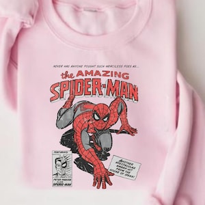 K&ouml;nnte beinhalten: Hellrosa Sweatshirt mit einem Comic-Grafik von Spider-Man. Das Design enth&auml;lt den Text "the AMAZING SPIDER-MAN" und andere Comic-Elemente. Das Sweatshirt ist aus weichem Stoff gefertigt.