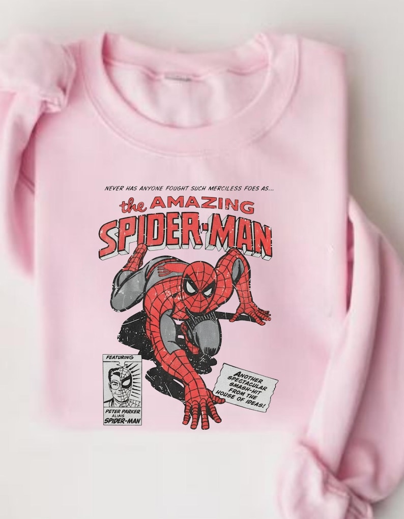 K&ouml;nnte beinhalten: Hellrosa Sweatshirt mit einem Comic-Grafik von Spider-Man. Das Design enth&auml;lt den Text "the AMAZING SPIDER-MAN" und andere Comic-Elemente. Das Sweatshirt ist aus weichem Stoff gefertigt.