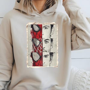 Op de afbeelding: Beige hoodie met een vintage afbeelding van Spider-Man's masker en de gezichten van drie acteurs. De afbeelding gebruikt rood, wit en zwart.