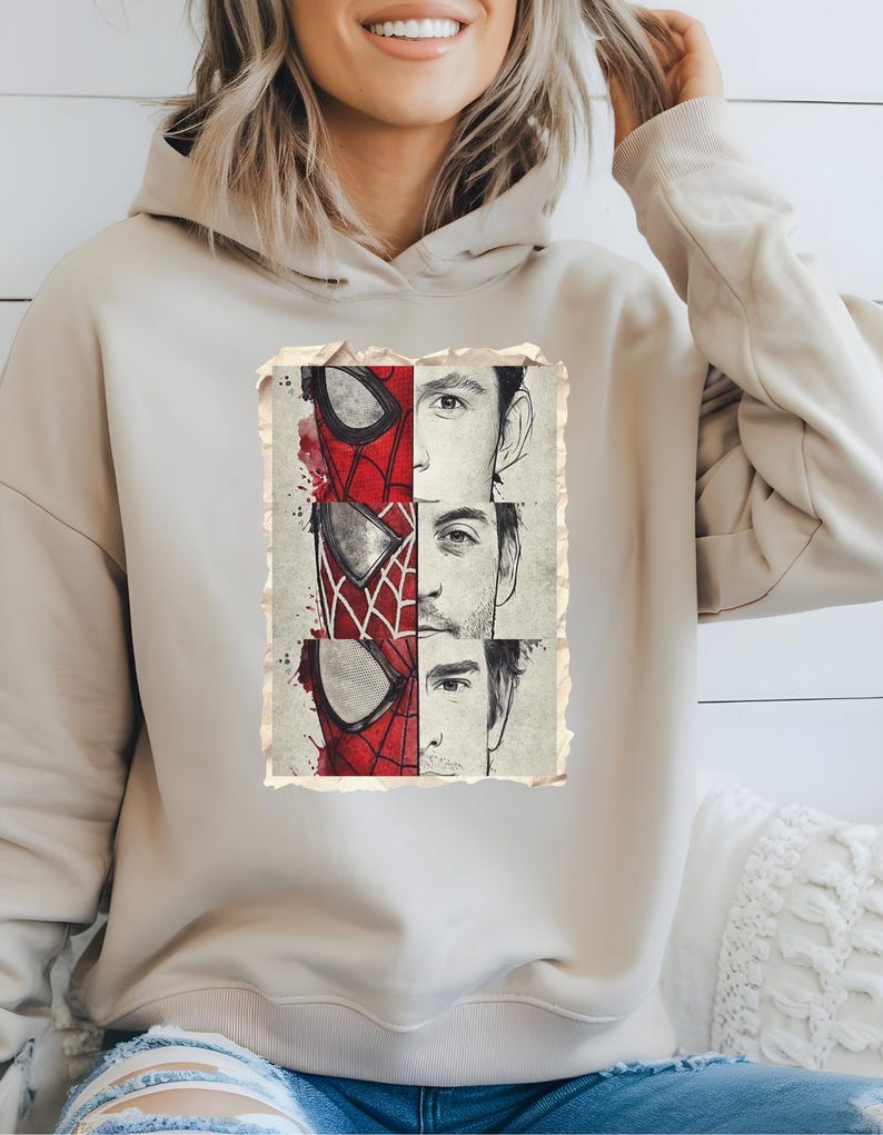 Op de afbeelding: Beige hoodie met een vintage afbeelding van Spider-Man's masker en de gezichten van drie acteurs. De afbeelding gebruikt rood, wit en zwart.