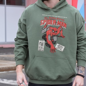 K&ouml;nnte beinhalten: Milit&auml;rgr&uuml;ner Hoodie mit einem Vintage-Comic-Grafik von Spider-Man. Das Design zeigt den Text "The Amazing Spider-Man" in Rot und Wei&szlig;, zusammen mit anderem Comic-Text. Der Hoodie hat eine K&auml;ngurutasche und eine Kapuze mit Kordelzug.