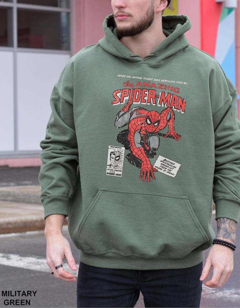 K&ouml;nnte beinhalten: Milit&auml;rgr&uuml;ner Hoodie mit einem Vintage-Comic-Grafik von Spider-Man. Das Design zeigt den Text "The Amazing Spider-Man" in Rot und Wei&szlig;, zusammen mit anderem Comic-Text. Der Hoodie hat eine K&auml;ngurutasche und eine Kapuze mit Kordelzug.
