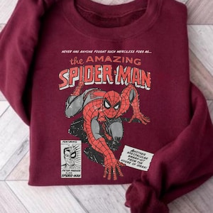 K&ouml;nnte beinhalten: Burgunderfarbenes Sweatshirt mit einem Vintage-Comic-Stil-Grafik von Spider-Man. Das Design enth&auml;lt den Text "the AMAZING SPIDER-MAN" und Textfelder mit "PETER PARKER" und "HOUSE OF IDEAS!"