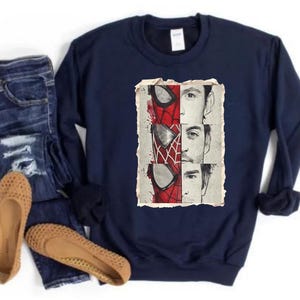 Op de afbeelding: Marineblauwe sweatshirt met een afbeelding van Spider-Mans masker en gezichten van acteurs die het personage speelden. De afbeelding is in rood, wit en zwart. De sweatshirt wordt gecombineerd met een blauwe jeans en beige ballerina's.
