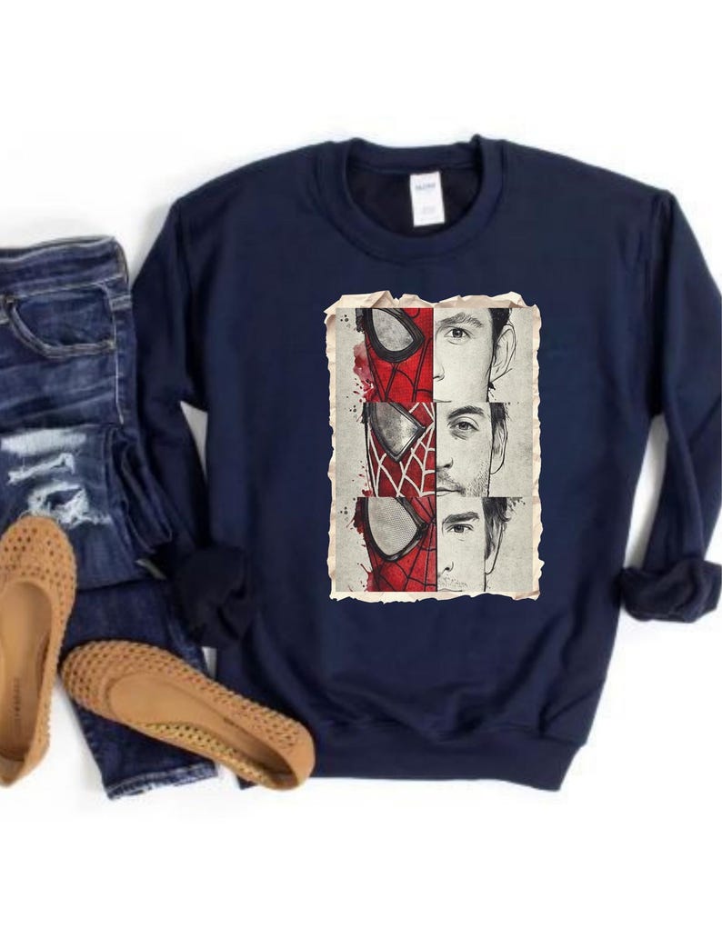 Op de afbeelding: Marineblauwe sweatshirt met een afbeelding van Spider-Mans masker en gezichten van acteurs die het personage speelden. De afbeelding is in rood, wit en zwart. De sweatshirt wordt gecombineerd met een blauwe jeans en beige ballerina's.