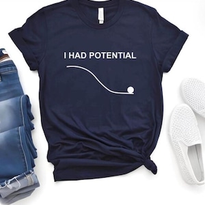 May include: Navy blue t-shirt with the text "I HAD POTENTIAL" above a white graphic of a ball rolling down a curved line. The shirt is styled with blue jeans and white slip-on shoes.
