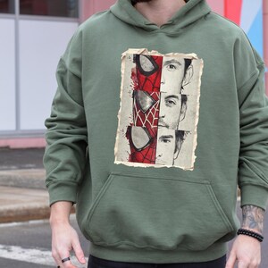Op de afbeelding: Militairgroene hoodie met een grafische print van Spider-Man. De print staat op een verweerde achtergrond. De hoodie heeft een kangoeroezak en een capuchon met trekkoord. De tekst "MILITARY GREEN" staat onderaan.