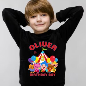 Puede incluir: Sudadera negra con el texto "Oliver the Birthday Boy" en rojo y amarillo, una carpa de circo y personajes de dibujos animados. El dise&ntilde;o incluye globos y el n&uacute;mero 5.