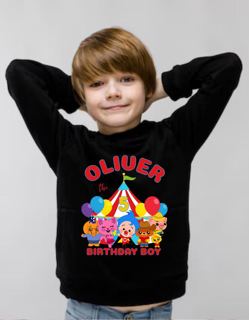 Puede incluir: Sudadera negra con el texto "Oliver the Birthday Boy" en rojo y amarillo, una carpa de circo y personajes de dibujos animados. El dise&ntilde;o incluye globos y el n&uacute;mero 5.