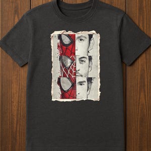 Op de afbeelding: Donkergrijs t-shirt met een afbeelding van Spider-Mans masker en de gezichten van drie acteurs. Het ontwerp is verdeeld in secties, met het Spider-Man masker in rood en zwart, en de gezichten van de acteurs in zwart en wit.