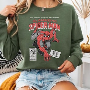 K&ouml;nnte beinhalten: Gr&uuml;nes Langarm-Sweatshirt mit einem Vintage-Comic-Grafik von Spider-Man. Die Grafik enth&auml;lt den Text "the AMAZING SPIDER-MAN" und andere Comic-Elemente. Das Sweatshirt wird mit Jeansshorts getragen.