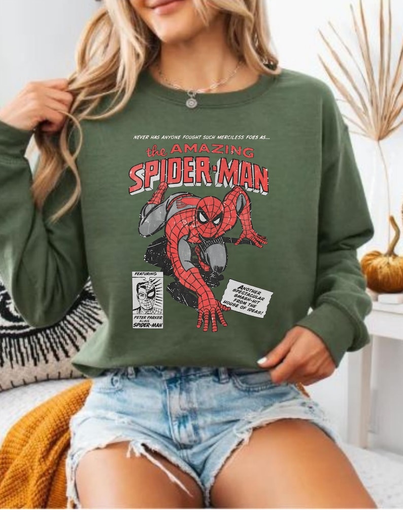 K&ouml;nnte beinhalten: Gr&uuml;nes Langarm-Sweatshirt mit einem Vintage-Comic-Grafik von Spider-Man. Die Grafik enth&auml;lt den Text "the AMAZING SPIDER-MAN" und andere Comic-Elemente. Das Sweatshirt wird mit Jeansshorts getragen.