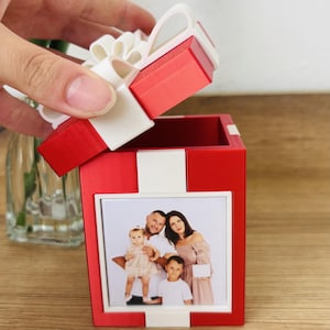 Pode incluir: Uma caixa de presente quadrada vermelha com tampa branca e um laço branco no topo. A frente da caixa apresenta uma inserção de foto quadrada exibindo um retrato de família de dois adultos e duas crianças. A caixa tem acabamento fosco.