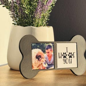 May include: A bone-shaped gray frame featuring two square inserts. The left insert displays a photograph of a person with a dog, while the right insert features the black text I WOOF YOU with two paw prints replacing the letter O.