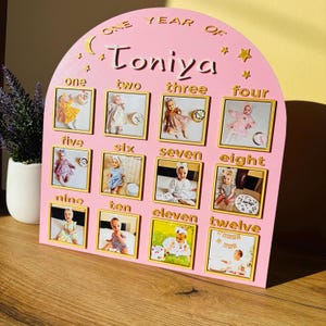 Peut inclure: Un panneau de suivi de croissance en bois rose en forme d'arche intitulé One Year of Toniya, avec douze cadres photo carrés étiquetés de un à douze en lettres dorées. Le panneau comporte des découpes dorées en forme de lune et d'étoiles.