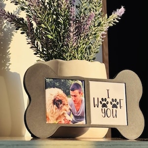 May include: A gray, bone-shaped picture frame with two openings. One holds a photo of a person and a dog. The other has the text "I woof you" with paw prints. A white vase with purple flowers is in the background.