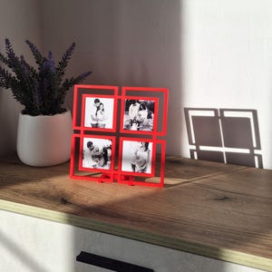 May include: A vibrant red photo frame, featuring four square apertures, each displaying a black and white photograph. The frame is positioned on a wooden surface, with a potted plant in the background. The frame's design is modern, suitable for family photos.