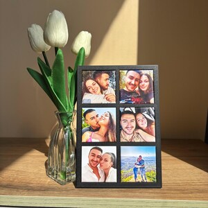 Può includere: Una cornice nera con sei slot per foto individuali, ciascuno riempito con una diversa fotografia a colori. La cornice è esposta su una superficie di legno accanto a un vaso di tulipani bianchi.