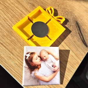 Può includere: Un supporto magnetico quadrato giallo con un magnete circolare nero e un piccolo fiocco giallo. Sotto c'è un magnete quadrato con una foto di una ragazza e un bambino. La foto è su una superficie di legno.