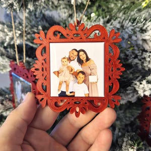 Può includere: Un ornamento natalizio quadrato rosso e ornato con una foto di famiglia. L'ornamento è appeso a un albero di Natale innevato. La foto mostra una famiglia di quattro persone. L'ornamento è tenuto da una mano.