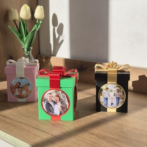 May include: Three square gift-shaped boxes with bows on top, each featuring a circular photo frame on the front. The boxes are pink with a white bow, green with a red bow, and black with a gold bow, displaying family photographs in the center.