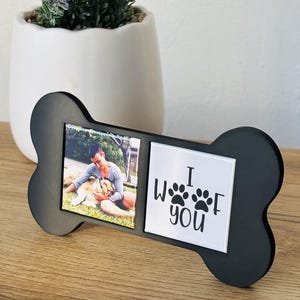 May include: Black bone-shaped photo frame with two sections. One section holds a photo of a person with a dog. The other section has the text "I Woof You" with paw prints. The frame is on a wooden surface.