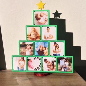 Może przedstawiać: Zielona ramka na zdjęcia w kształcie piramidy z żółtą gwiazdką na szczycie. Ramka mieści dziesięć kwadratowych fotografii przedstawiających różne portrety rodzinne i zdjęcia niemowląt, ułożonych w cztery poziome rzędy o rosnącej szerokości.