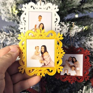 Könnte beinhalten: Weihnachtsbaumschmuck mit gerahmten Familienfotos. Die Rahmen sind gold, weiß und rot mit kunstvollen Designs. Ein goldener Rahmen, der von einer Hand gehalten wird, zeigt ein Foto einer Frau und eines Kindes.
