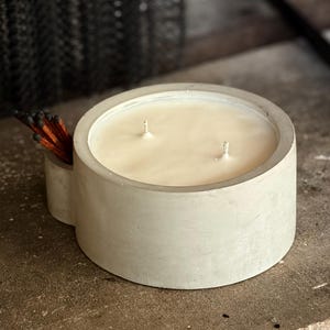 May include: A round, light gray concrete candle with three wicks and a creamy wax fill. A small, attached compartment holds a bundle of matches. The candle sits on a rough, gray surface.