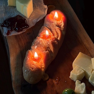May include: A lit candle shaped like a loaf of bread with three flames, placed on a wooden serving board. Cubes of cheese, butter, and olives surround the candle, with a small dish of jam. The candle's warm glow contrasts with the dark background.