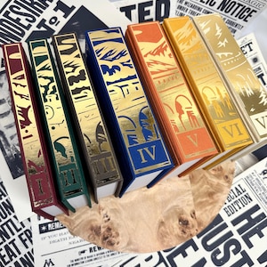 May include: A collection of hardcover books with gold foil accents and spines in various colors, including burgundy, green, blue, orange, and yellow. The books are arranged with related ephemera, including posters with text such as "Undesirable No. 1" and "Ministry of Magic Public Notice."