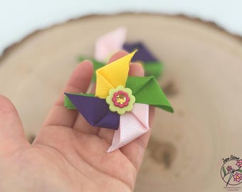 May include: A colorful hair bow in a pinwheel design. The bow features yellow, green, purple, and pink ribbons, with a small flower embellishment at the center. The bow is held in a hand, with a blurred wooden background.