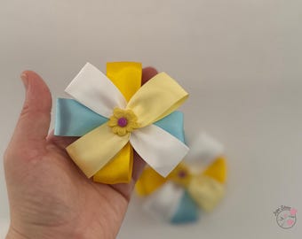May include: A yellow, white, and blue ribbon bow with a small purple flower in the center. The bow is tied in a knot and is ready to be used as a hair accessory.