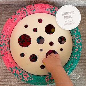 Könnte beinhalten: Ein kreisförmiges Holzspielzeug mit einem cremefarbenen Mittelteil mit unterschiedlich großen Löchern. Der Rand ist rosa und grün. Der Text auf dem Bild lautet "STAPELSTEIN EINLAGE WAS IST GROB UND WAS KLEIN?"