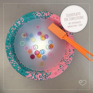 Könnte beinhalten: Ein farbenfrohes Spielzeugset mit einer gesprenkelten Schüssel in Rosa und Türkis und einem durchscheinenden Zentrum. Das Set enthält ein orangefarbenes Werkzeug und kleine, farbige Scheiben. Der Text lautet "LEUCHTPLATTE FÜR STAPELSTEINE" und "MIT OPTIONALEN MAGNETSTAB + CHIPS".