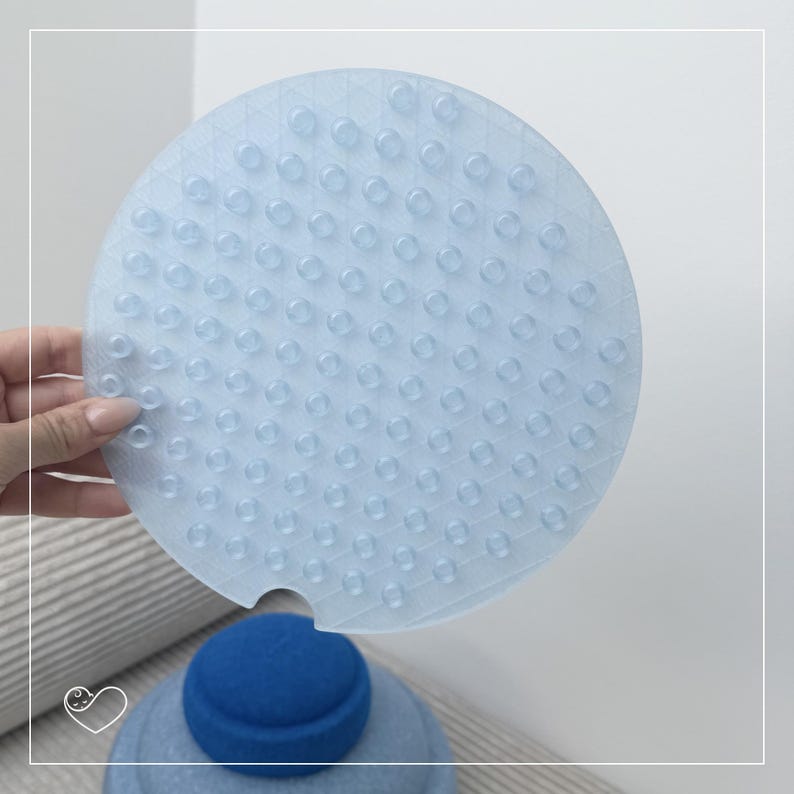 May include: A light blue, round bath mat with numerous small, raised bumps for grip. The mat is held above a blue object, possibly a bath toy or accessory. The design suggests a focus on safety and functionality in a bathroom setting.