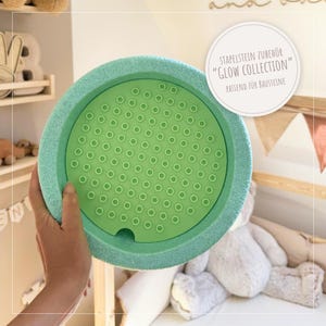 Könnte beinhalten: Ein rundes Spielzeugzubehör in zwei Grüntönen. Der äußere Ring ist ein strukturiertes, helles Türkis, während die innere Oberfläche ein glattes, helles Grün mit zahlreichen kleinen, kreisförmigen Vertiefungen aufweist. Text auf dem Bild lautet "STAPELSTEIN ZUBEHÖR 'GLOW COLLECTION' PASSEND FÜR BAUSTEINE."