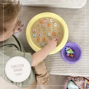 Könnte beinhalten: Ein gelbes Alphabet-Puzzle aus Holz mit bunten Buchstaben. Das Puzzle ist rund und hat eine Holzbasis mit Buchstabenausschnitten. Die Hand eines Kindes zeigt auf einen Buchstaben. Eine lila Schüssel mit zusätzlichen Buchstaben befindet sich neben dem Puzzle.