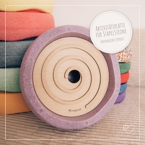 Könnte beinhalten: Ein kreisförmiges, lavendelfarbenes Aktivitätsbrett mit einem hölzernen Spiraleinsatz. Das Brett hat einen erhöhten Rand und der Text "AKTIVITÄTSPLATTE FÜR STAPELSTEINE UNENDLICHE SPIRALE" ist sichtbar. Bunte, gestapelte Schaumstoffblöcke sind im Hintergrund.