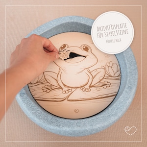 Könnte beinhalten: Eine Holz-Aktivitätsplatte mit einer Frosch-Illustration, entworfen für Stapelsteine. Die Platte hat einen hellblauen Rand und den Text "AKTIVITÄTSPLATTE FÜR STAPELSTEINE FÜTTERE MICH". Eine Hand platziert eine Holzblume im Maul des Frosches.