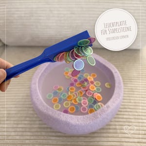 Könnte beinhalten: Ein blauer Magnetstab hebt farbige, durchscheinende Scheiben aus einer lavendelfarbenen Schale. Die Scheiben sind in verschiedenen Farben, darunter Rosa, Grün, Orange und Gelb. Die Schale steht auf einer hellgrauen Oberfläche. Text auf einem weißen Kreis lautet "LEUCHTPLATTE FÜR STAPELSTEINE SPIELERISCH LERNEN".