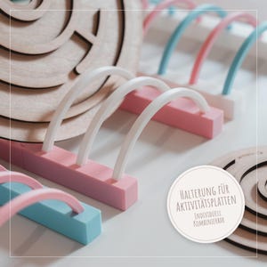 Könnte beinhalten: Nahaufnahme von farbenfrohen Holz- und Kunststoffkomponenten für Aktivitätstafeln. Die Komponenten umfassen rosa, blaue und weiße Bögen sowie kreisförmige Holzteile. Der Text auf dem Bild lautet "Halterung für Aktivitätsplatten Individuell Kombinierbar."