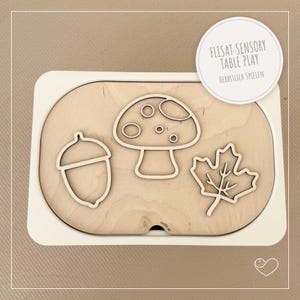 Könnte beinhalten: Ein Holz-Sensorik-Tischspielset mit weißem Rahmen. Das Set enthält eine Holzbasis mit Ausschnitten von Eichel, Pilz und Blatt. Der Text "FLISAT SENSORY TABLE PLAY" befindet sich auf einem weißen kreisförmigen Etikett.