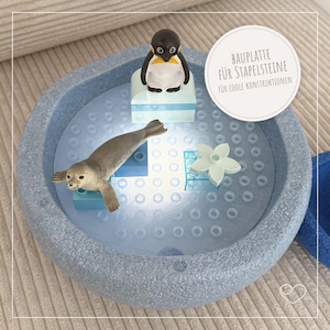 Könnte beinhalten: Eine hellblaue, strukturierte Spielbasis mit einer Pinguin- und Robbenfigur, zusammen mit einer weißen Blume und blauen Bausteinen. Der Text auf dem Bild lautet "BAUPLATTE FÜR STAPELSTEINE FÜR COOLE KONSTRUKTIONEN".
