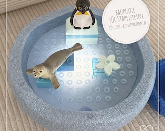 May include: A light blue, textured play base with a penguin and seal figurine, along with a white flower and blue building blocks. The text on the image reads "BAUPLATTE FÜR STAPELSTEINE FÜR COOLE KONSTRUKTIONEN".