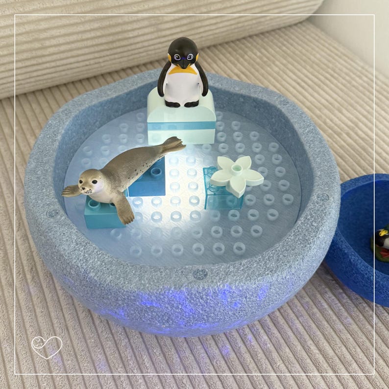 May include: A light blue, textured, round toy with a penguin and seal figurine inside. The toy has a light blue base with a series of small holes. The penguin is black and white with a yellow beak. The seal is gray.