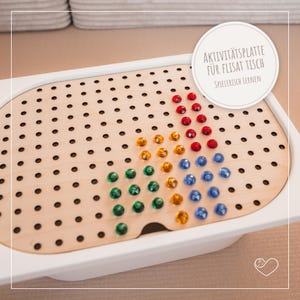 Könnte beinhalten: Aktivitätsplatte aus Holz mit weißem Rahmen, mit einer perforierten Oberfläche und bunten, edelsteinartigen Stiften, die in einem Regenbogenmuster angeordnet sind. Das Brett enthält den Text "AKTIVITÄTSPLATTE FÜR FLISAT TISCH SPIELERISCH LERNEN" in einem weißen Kreis.